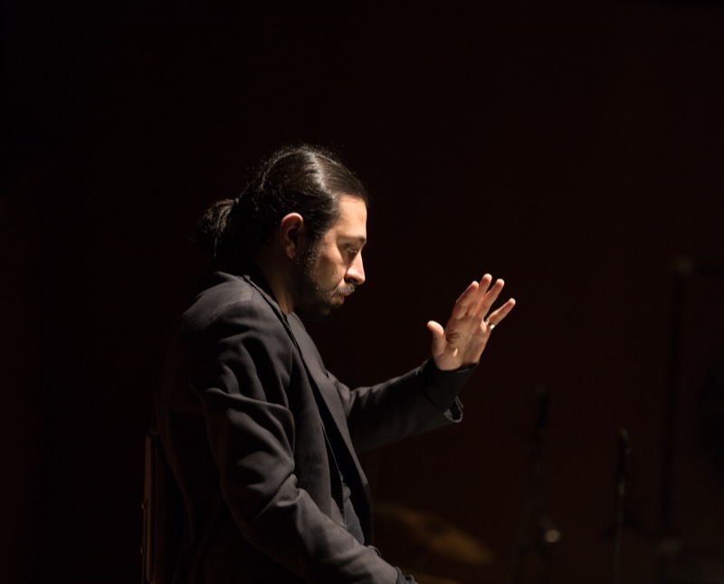 Francesco Bossaglia conducting mid-gesture in the pit at LAC Lugano, lit by a single warm rim light against a black background.