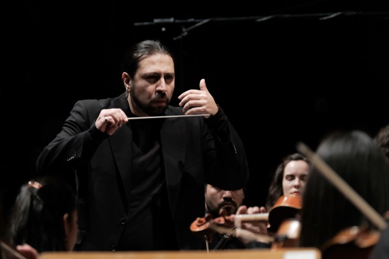 Francesco Bossaglia on a dark stage, baton raised mid-beat, strong side light, string section and bows visible in foreground.