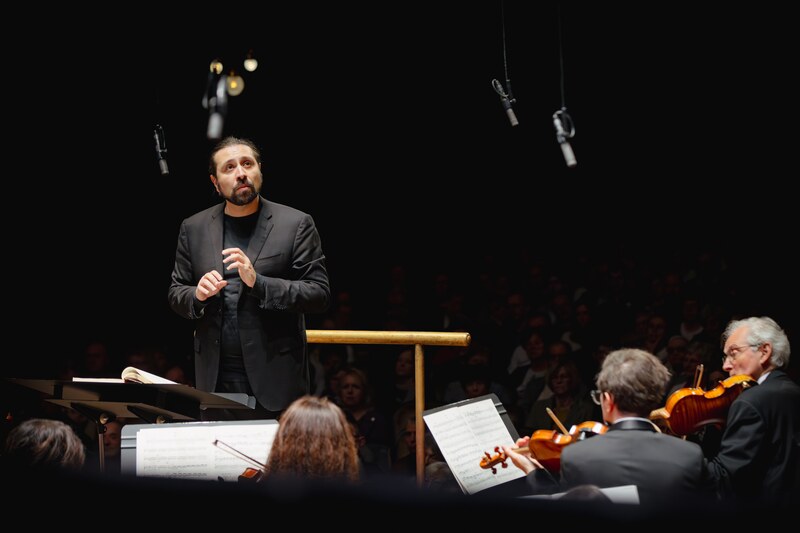 Full orchestra viewed from the hall, gold rail and packed audience in a grand auditorium.