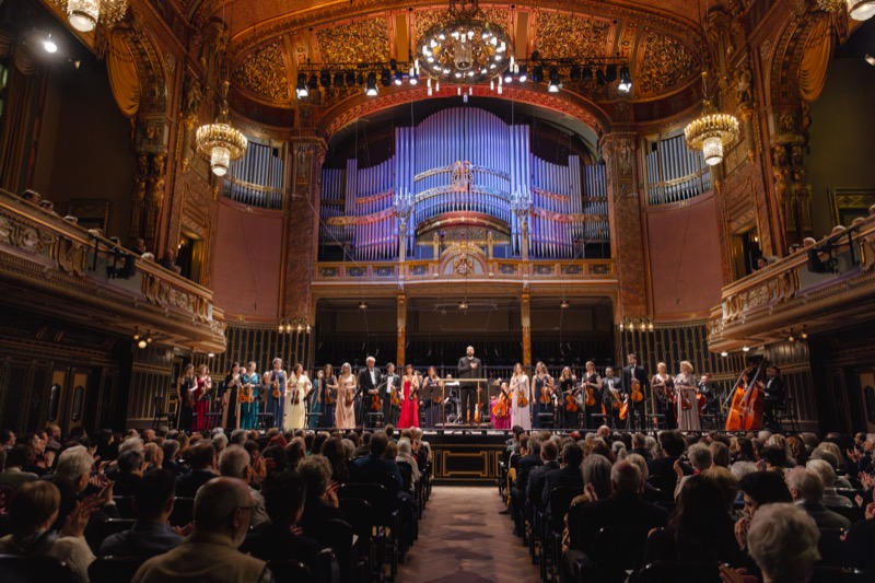 Wide shot of a gilded concert hall with the full ensemble lined up on stage for a curtain call, audience applauding.