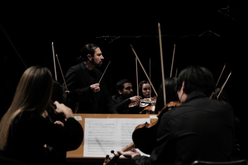 Francesco Bossaglia conducting in profile in a dark hall, orchestra bows raised in foreground, lit by a single hanging spotlight.
