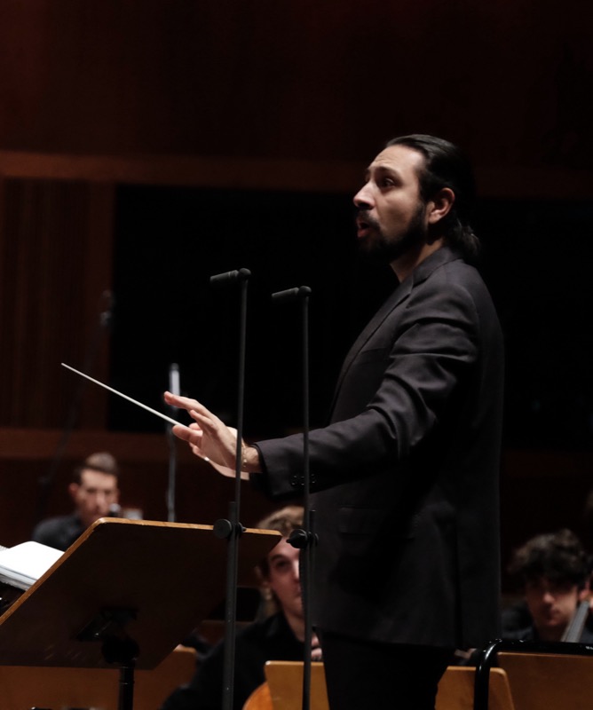 Francesco Bossaglia conducting on a dark stage, mouth open mid-phrase, baton hand raised.