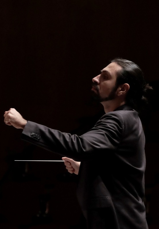 Francesco Bossaglia conducting, shot from behind in dark theatrical lighting, baton extended.