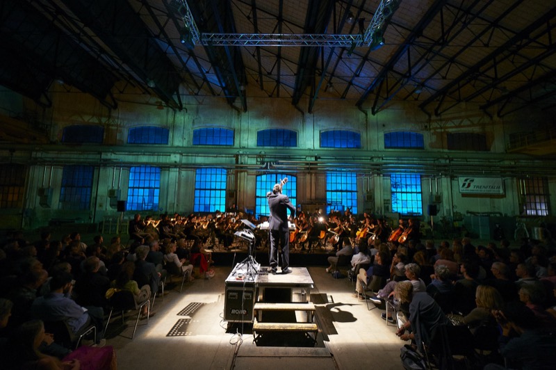 Francesco Bossaglia conducting from behind in a repurposed industrial warehouse under blue stage lighting, Transart 2016.